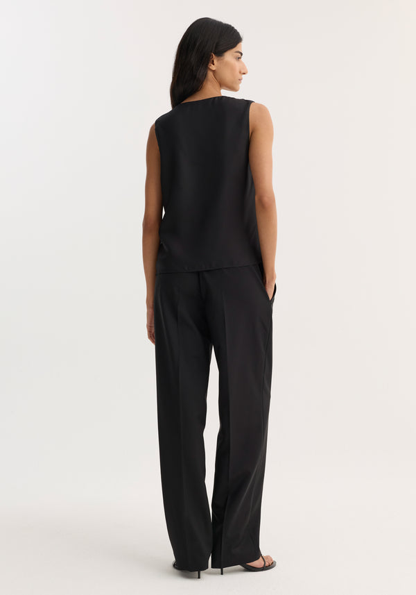 Rear-view of a woman wearing a black scoop-neck silk top, showcasing its loose fit and wide neckline, paired with matching wide-leg trousers and heeled sandals.