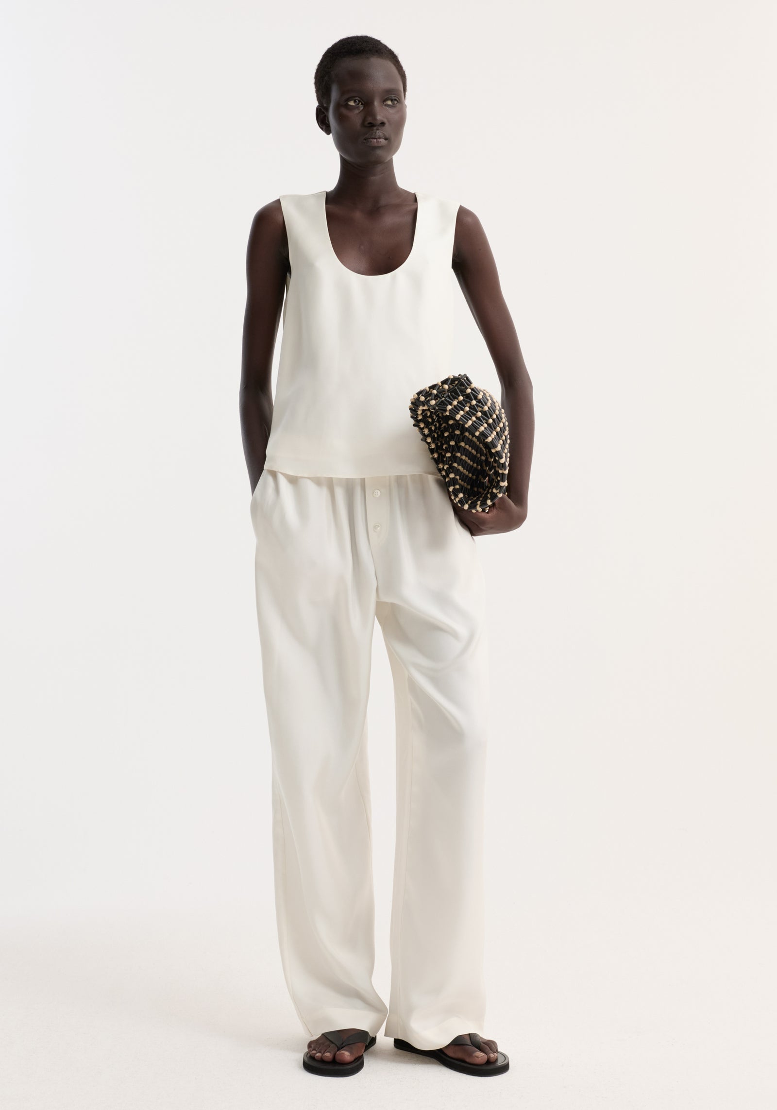 Model wearing a Scoop-neck silk top | cream, featuring a loose fit and wide scoop neck, paired with wide-leg trousers and holding a black woven clutch.