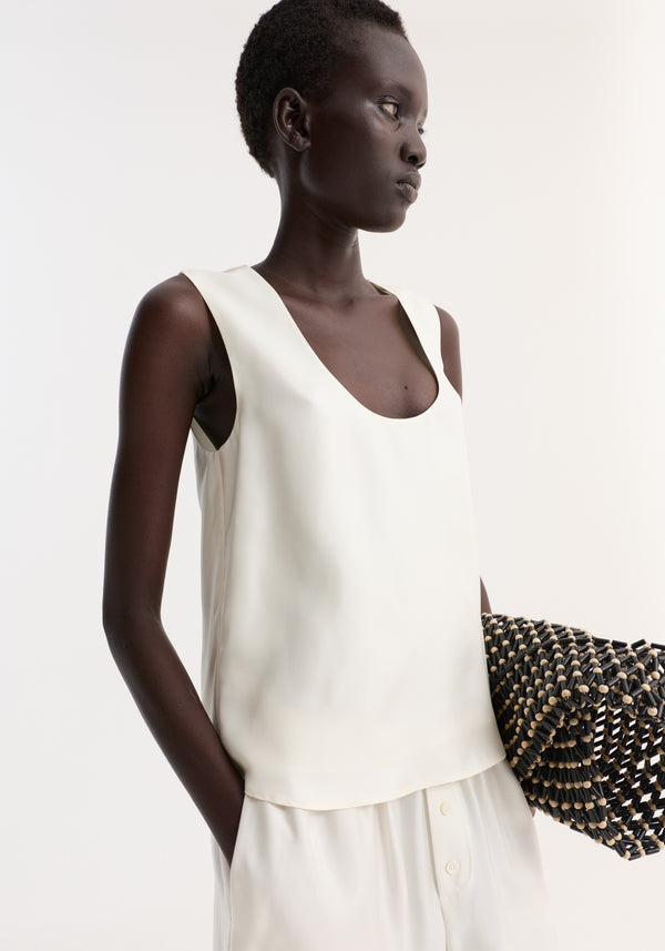 Model wearing scoop-neck silk top | cream, showcasing its wide scoop neck and loose fit, paired with matching trousers, holding a woven clutch.