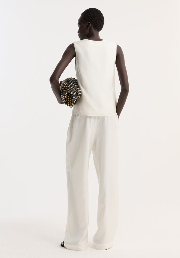 Woman in a scoop-neck silk top, cream, with loose fit, paired with wide-leg trousers, holding a beaded clutch and wearing black sandals.