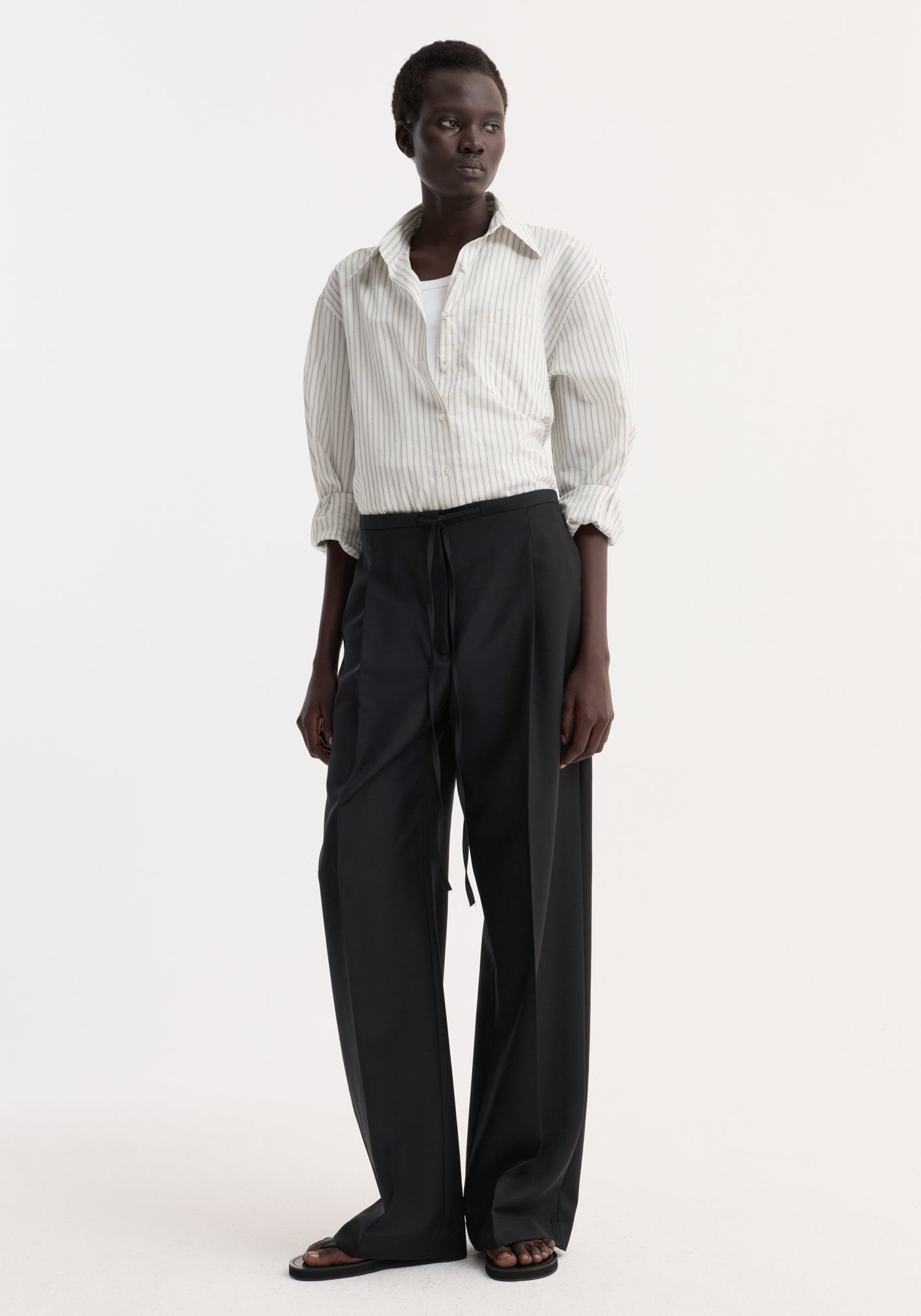 Dark-skinned woman wearing Relaxed drawstring trousers in black, featuring a wide leg and casual chic drawstring waistband, paired with a pin-striped shirt and black sandals.
