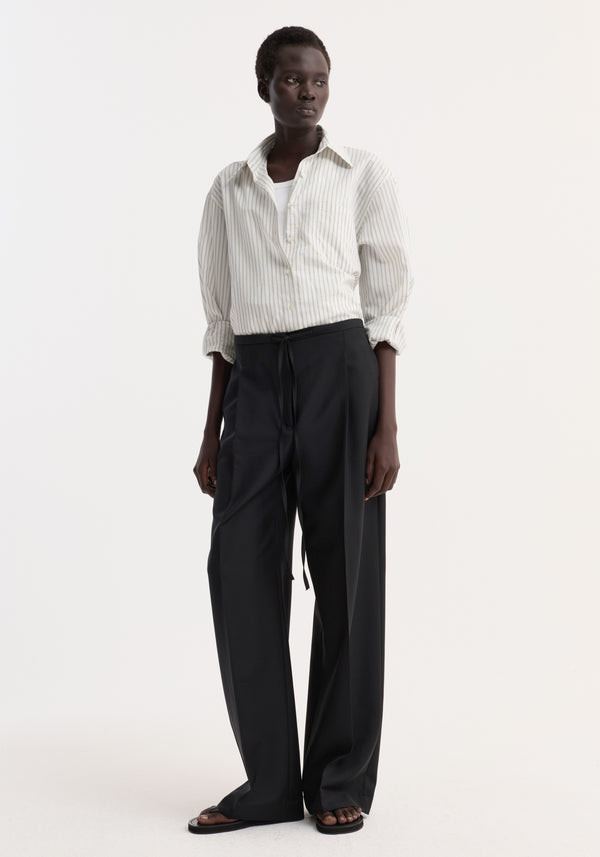 Dark-skinned woman wearing Relaxed drawstring trousers in black, featuring a wide leg and casual chic drawstring waistband, paired with a pin-striped shirt and black sandals.