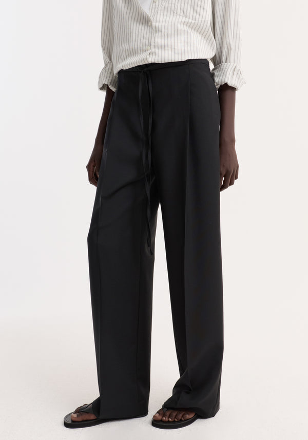 Relaxed drawstring trousers in black, loose-fit, wide-leg with drawstring waist, styled with rolled-sleeve shirt and sandals, showcasing a long, flowing silhouette.