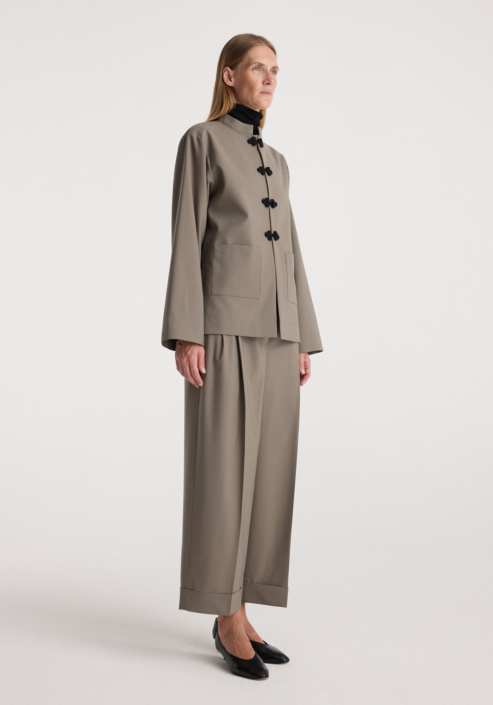 Woman wearing high-waist turn-up trousers | taupe paired with a matching taupe jacket and black pointed shoes, standing against a plain light background.
