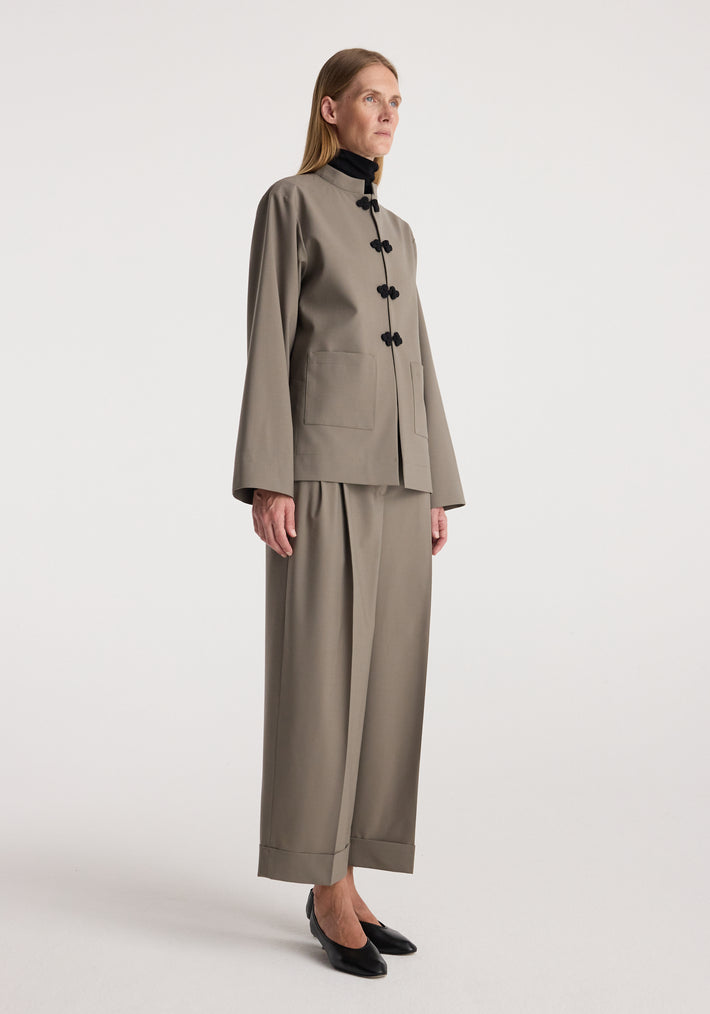 Woman wearing high-waist turn-up trousers | taupe paired with a matching taupe jacket and black pointed shoes, standing against a plain light background.