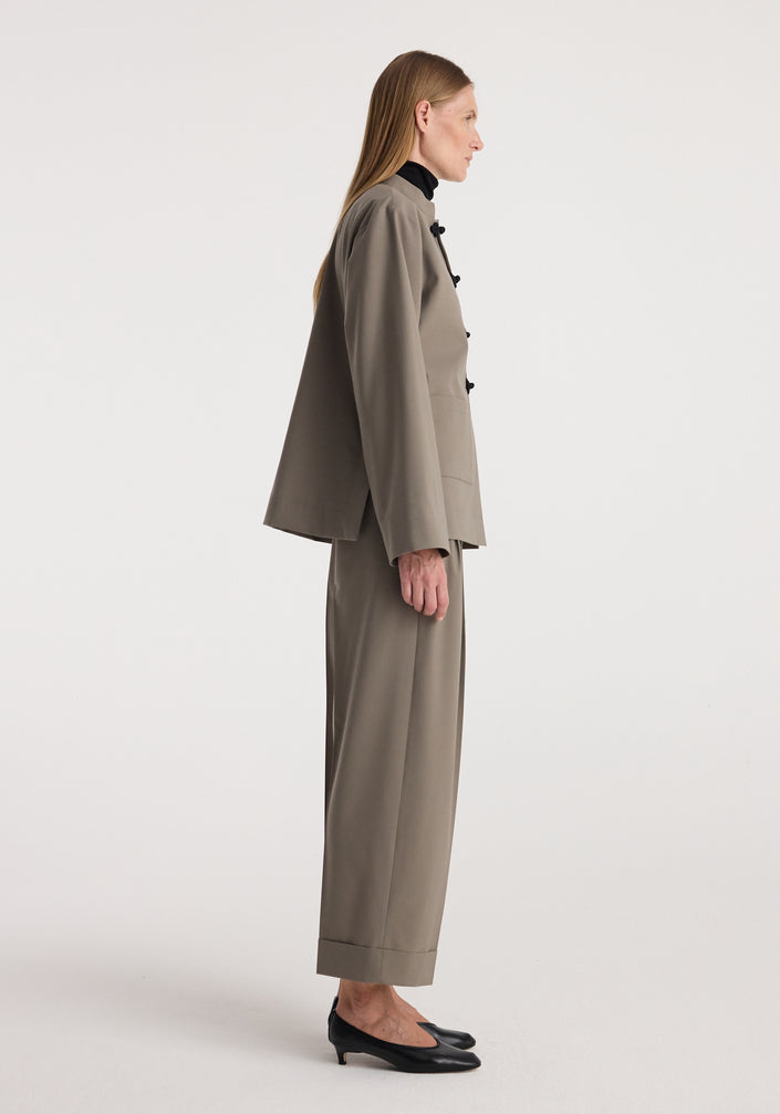 Woman wearing high-waist turn-up trousers | taupe paired with a matching taupe jacket and black pointed shoes, standing against a plain light background.