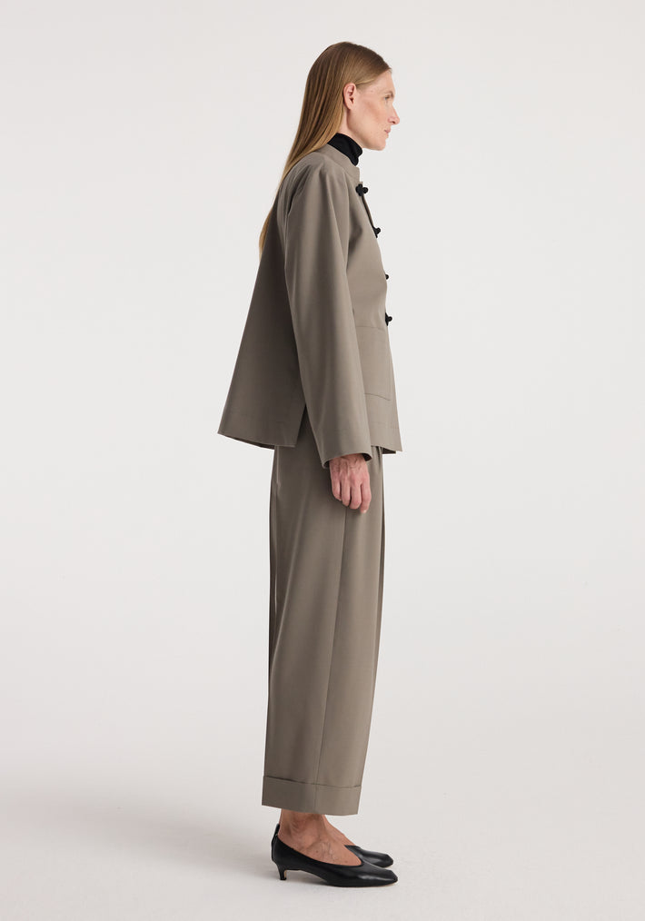 Side view of a woman wearing high-waist turn-up trousers | taupe paired with a matching taupe blazer, black top underneath, and black low-heeled shoes against a plain background.