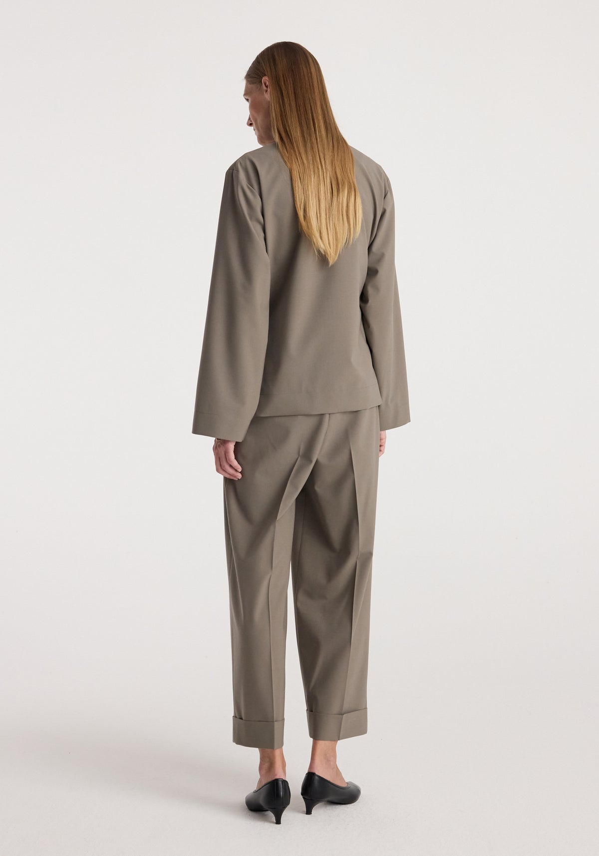 Woman wearing High-waist turn-up trousers | taupe with a matching long-sleeve top and black heeled shoes, standing against a plain light background showing the back view.