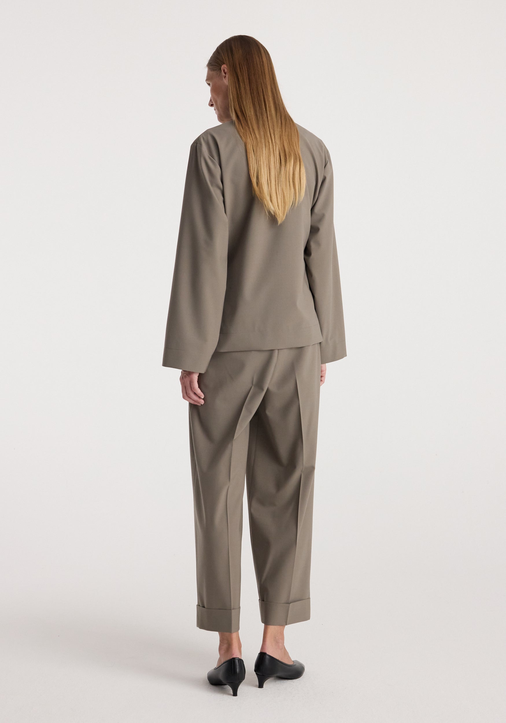 Woman wearing High-waist turn-up trousers | taupe with a matching long-sleeve top and black heeled shoes, standing against a plain light background showing the back view.