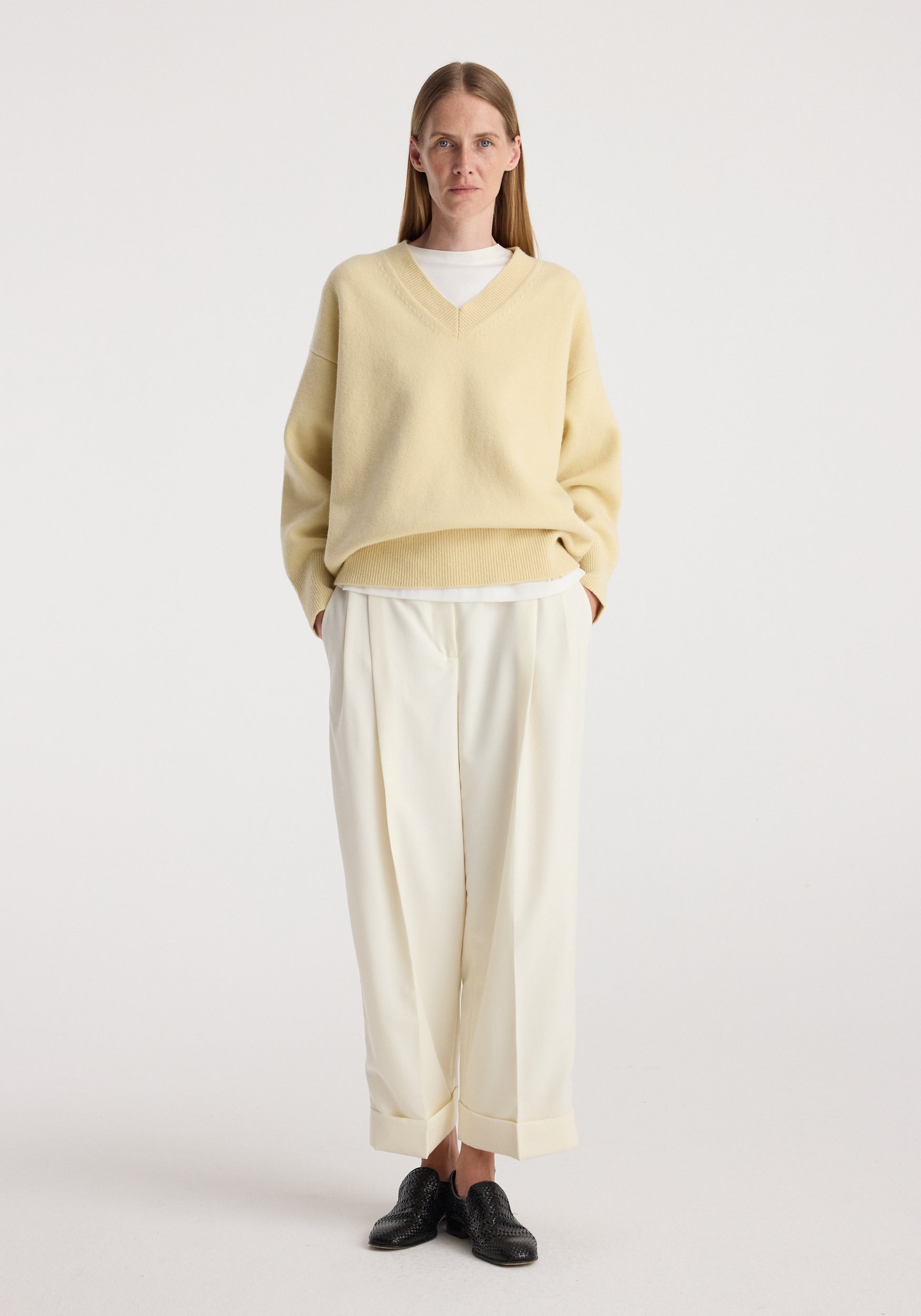 Woman standing wearing a light yellow V-neck sweater over a white top paired with High-waist turn up trousers | off white and black textured shoes against a plain white background.