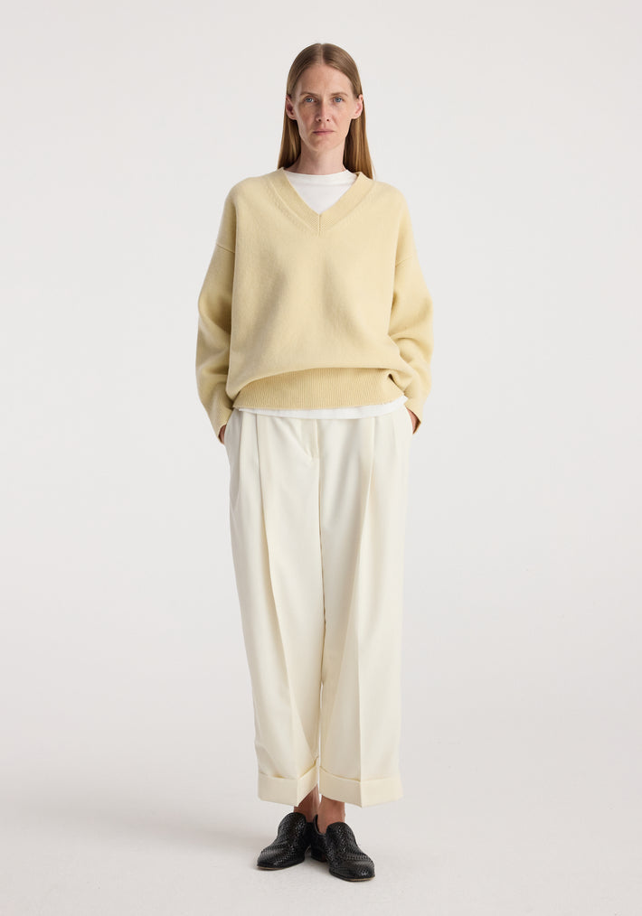 Woman standing wearing a light yellow V-neck sweater over a white top paired with High-waist turn up trousers | off white and black textured shoes against a plain white background.
