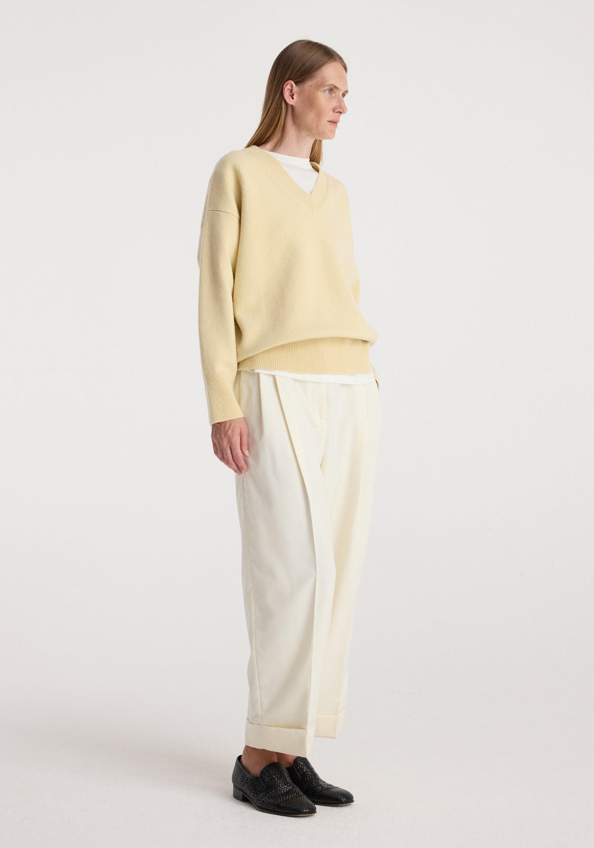 Woman wearing a light beige V-neck sweater and off white high-waist turn up trousers | off white paired with black loafers against a plain white background.