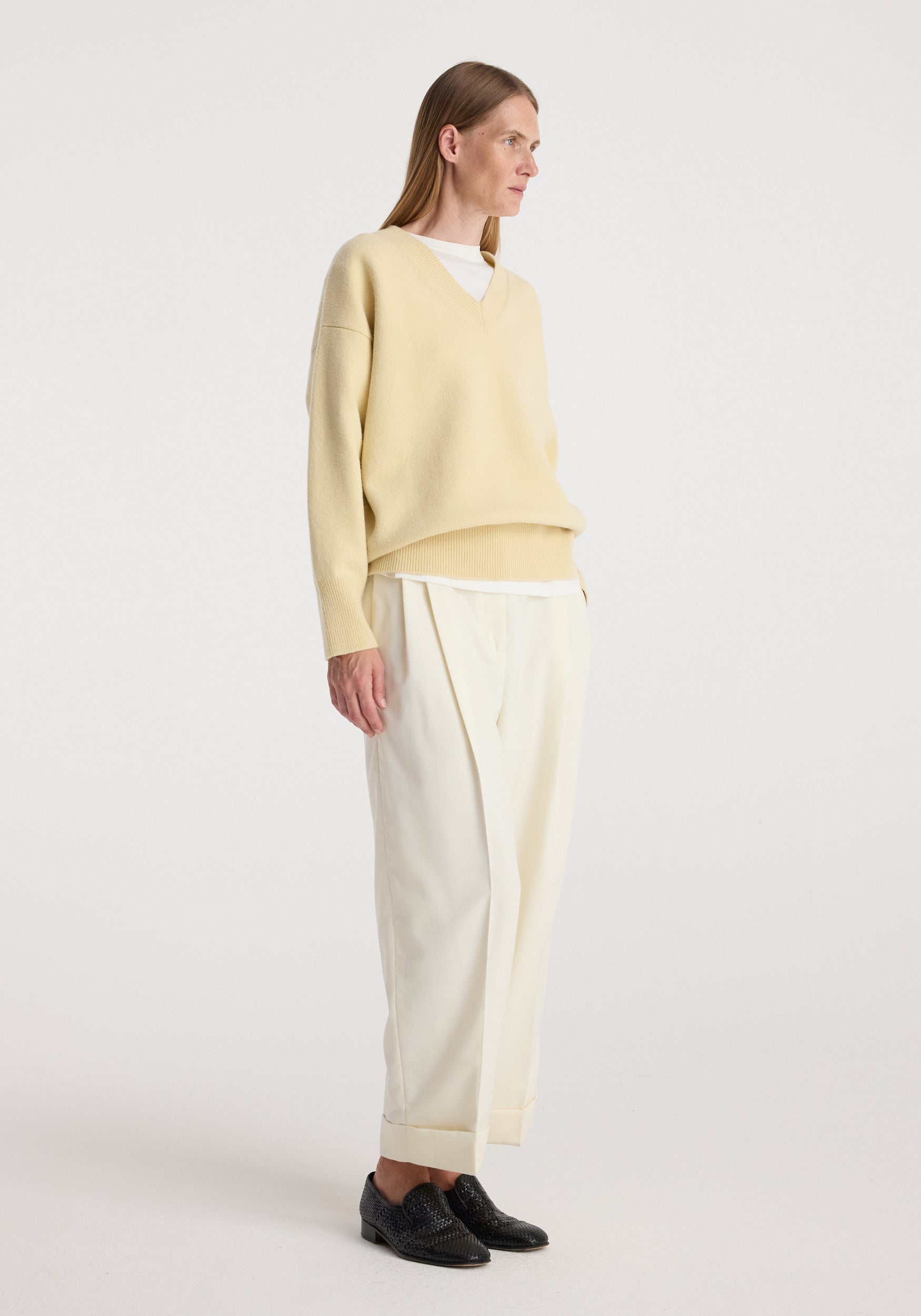 Woman wearing a light beige V-neck sweater and off white high-waist turn up trousers | off white paired with black loafers against a plain white background.