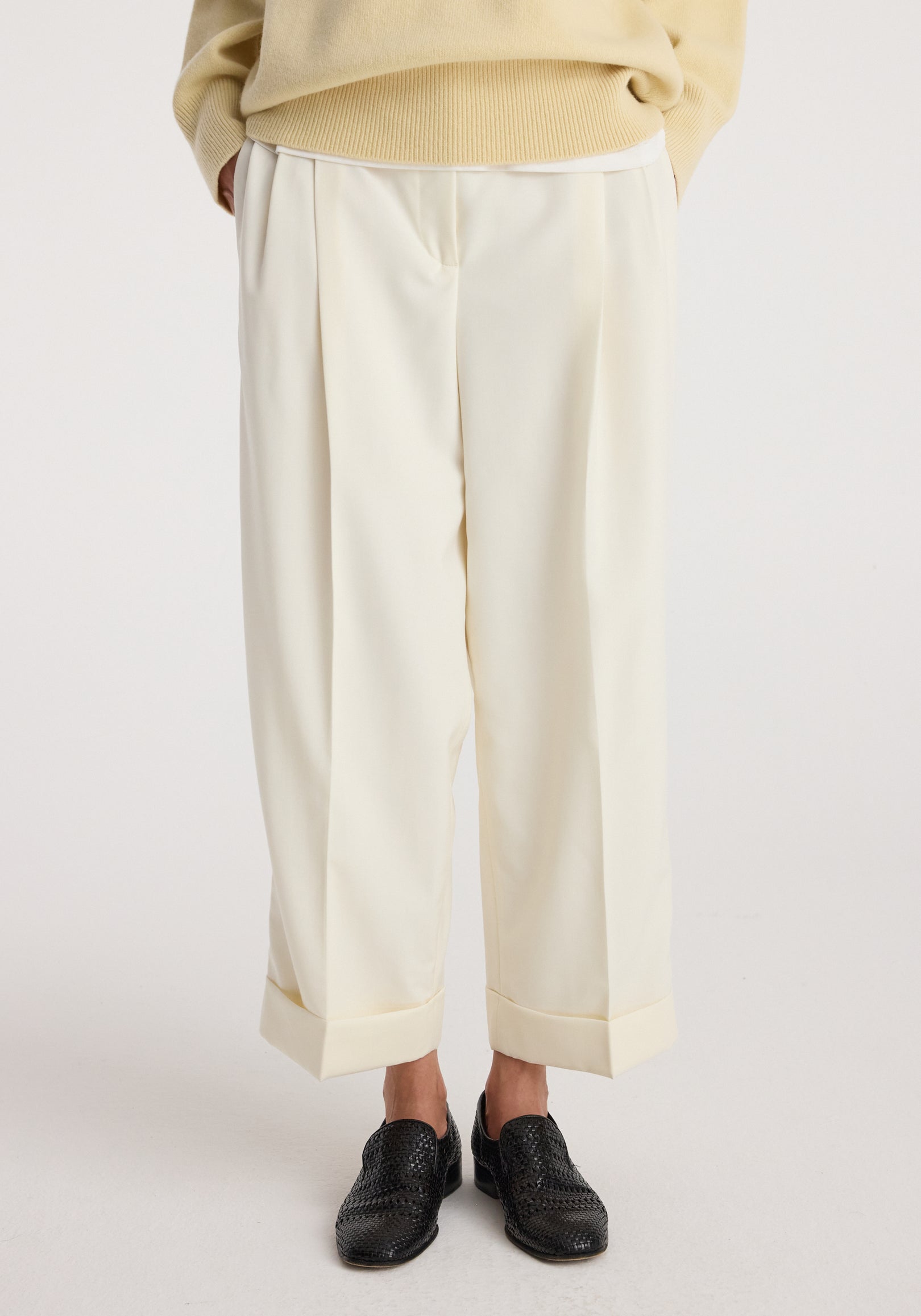 Close-up of a person wearing High-waist turn up trousers | off white paired with black woven shoes against a plain background.