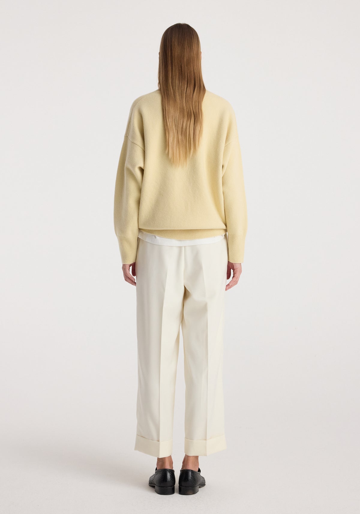 Woman wearing high-waist turn up trousers | off white with a loose beige sweater and black shoes, standing facing away on a plain background.