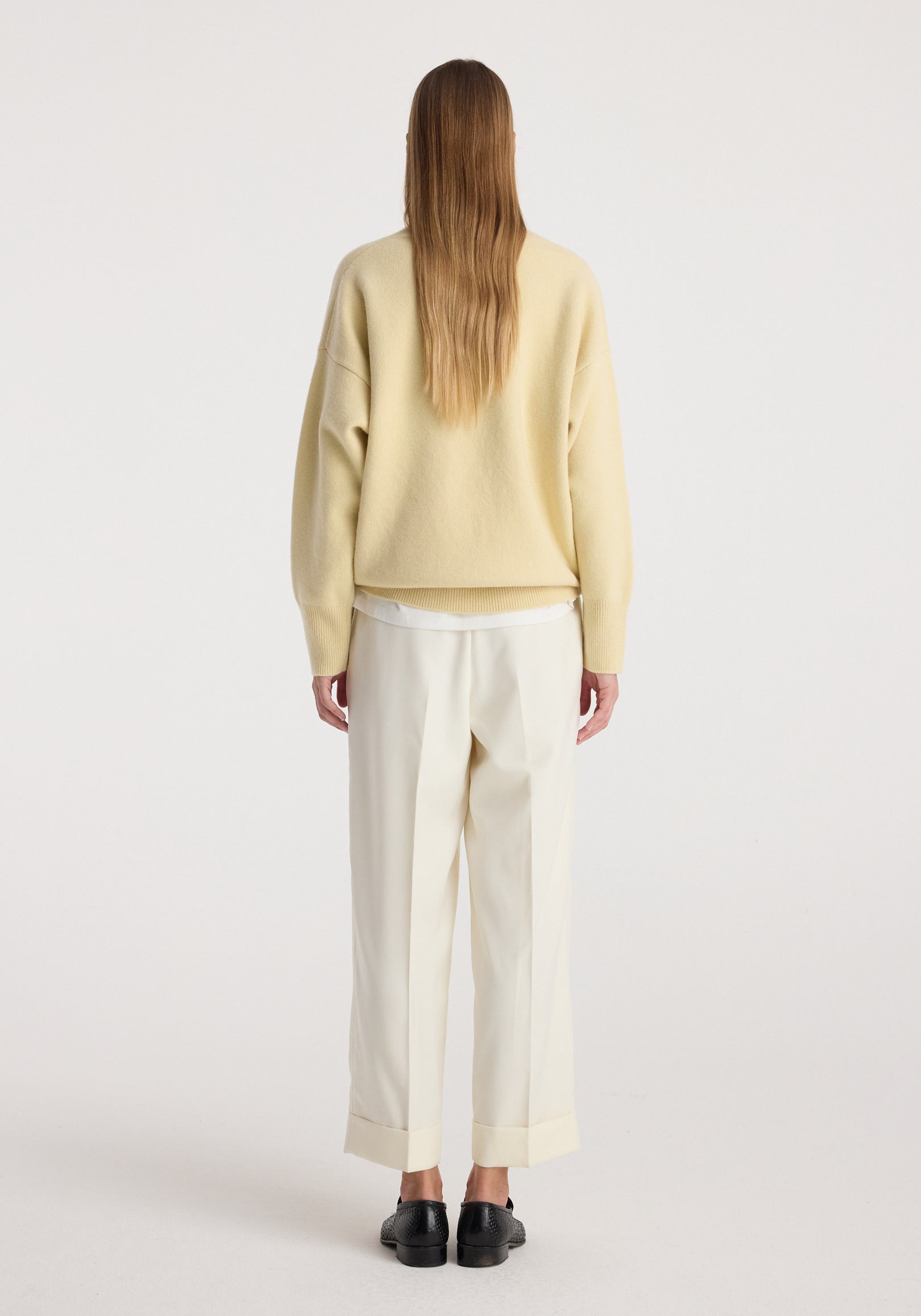 Woman wearing high-waist turn up trousers | off white with a loose beige sweater and black shoes, standing facing away on a plain background.