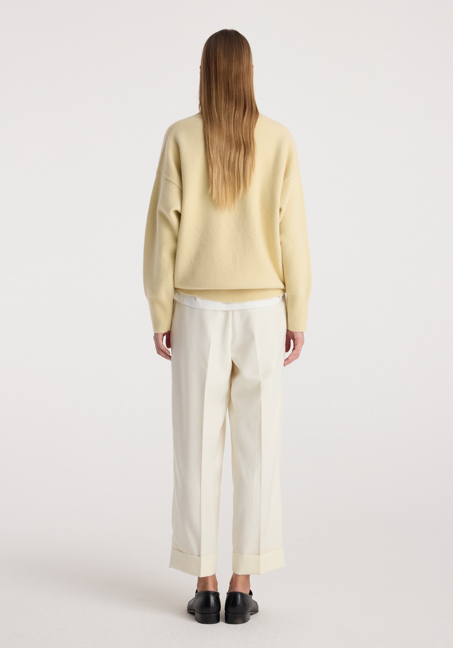 Woman wearing high-waist turn up trousers | off white with a loose beige sweater and black shoes, standing facing away on a plain background.