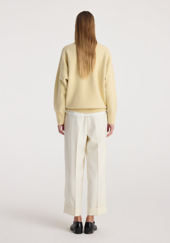 Woman wearing high-waist turn up trousers | off white with a loose beige sweater and black shoes, standing facing away on a plain background.