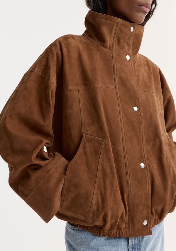 Person wearing the Reinvented suede jacket | cognac, featuring a high stand-up collar, snap-button closure, oversized pockets, and elasticized hem for a relaxed bomber fit.