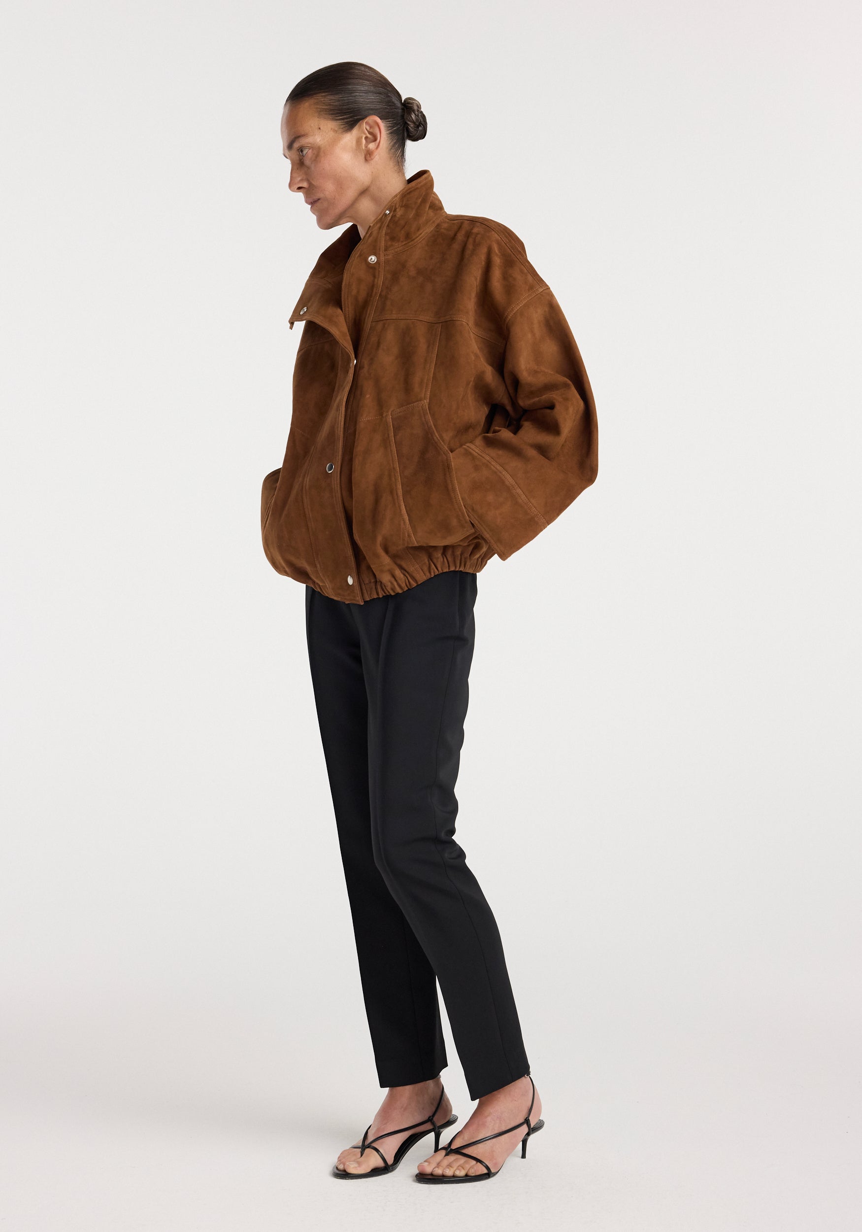 Side view of a woman wearing a loose-fitting Reinvented suede jacket | cognac in rich brown with black slim pants and strappy black heels against a plain white background.