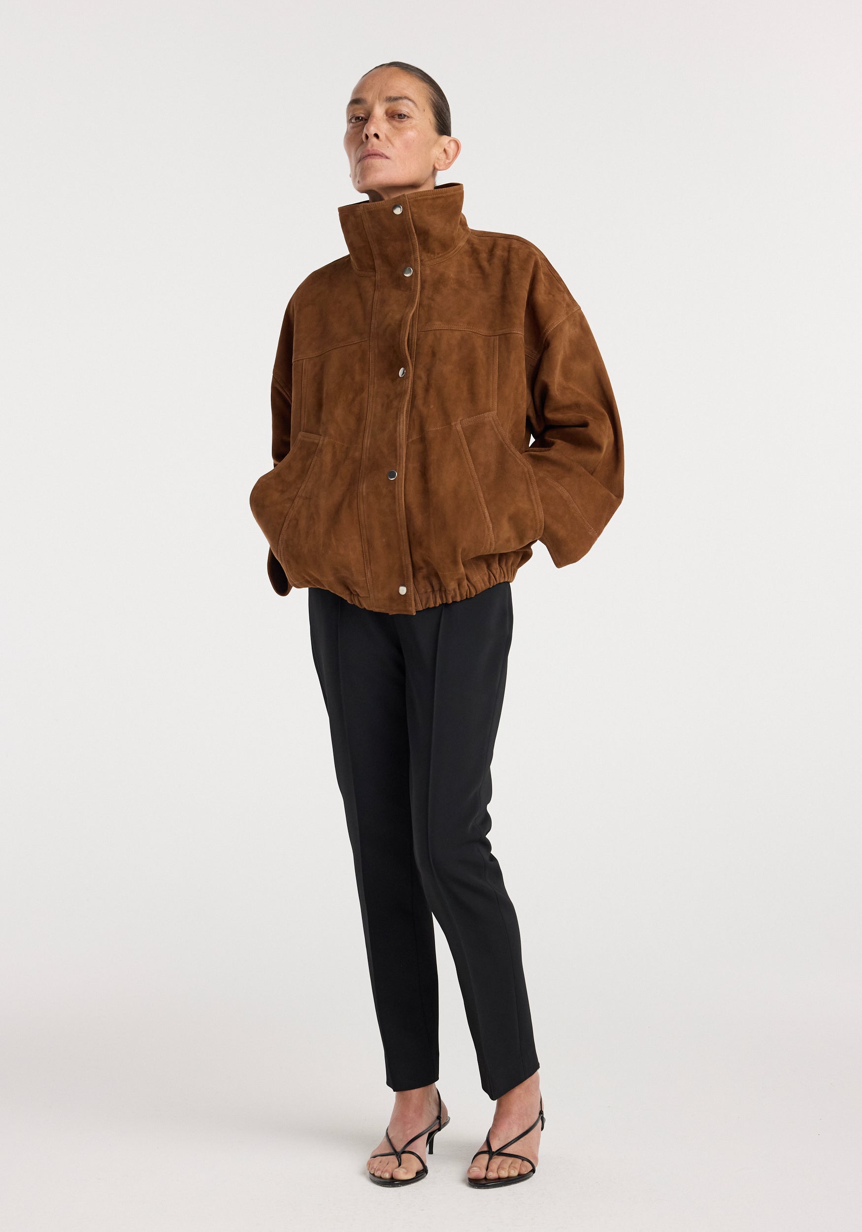 Woman wearing a cognac-colored Reinvented suede jacket | cognac with a high collar and snap buttons, styled with black tapered pants and strappy heels.