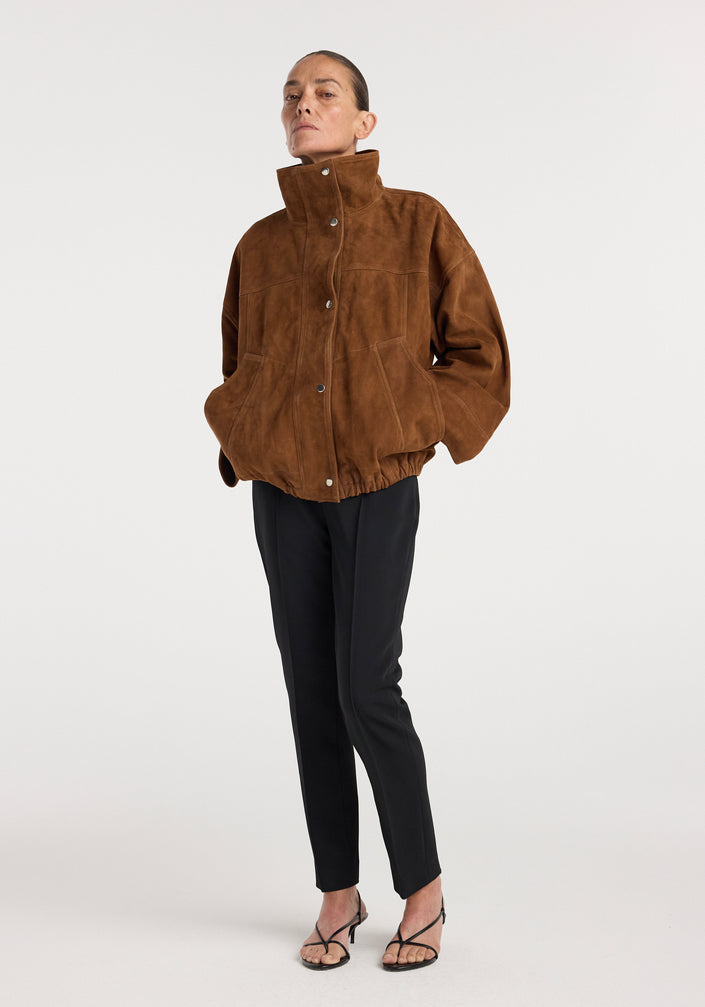 Woman wearing a cognac-colored Reinvented suede jacket | cognac with a high collar and snap buttons, styled with black tapered pants and strappy heels.