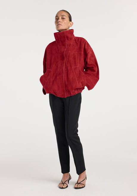 Woman wearing a high-collared red suede jacket with a relaxed fit and elastic hem, paired with slim black pants and strappy black heels, styled in a minimalist setting, showcasing the Reinvented suede jacket | red.
