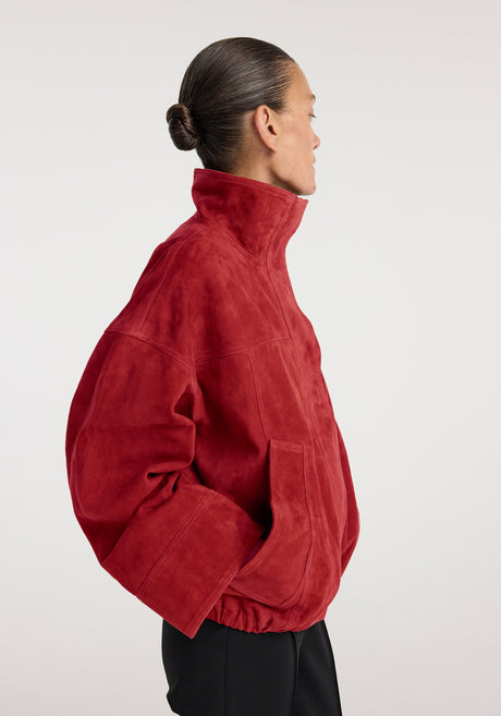 Woman wearing a high-collared red suede jacket with a relaxed fit and elastic hem, paired with slim black pants and strappy black heels, styled in a minimalist setting, showcasing the Reinvented suede jacket | red.