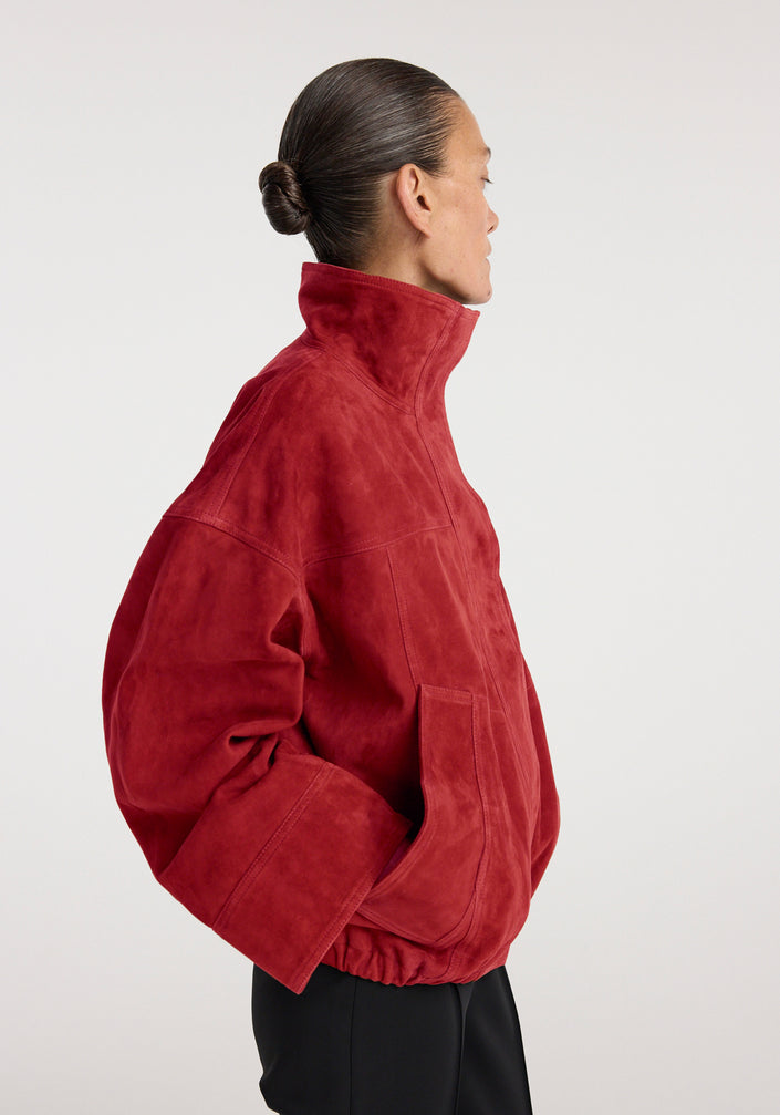 Woman wearing a high-collared red suede jacket with a relaxed fit and elastic hem, paired with slim black pants and strappy black heels, styled in a minimalist setting, showcasing the Reinvented suede jacket | red.