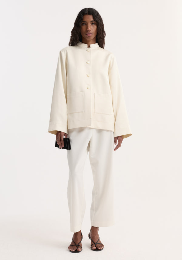 Model wearing a Pearl Button Satin Jacket in cream with a mandarin collar and front patch pockets, paired with matching straight-leg trousers and holding a black clutch.