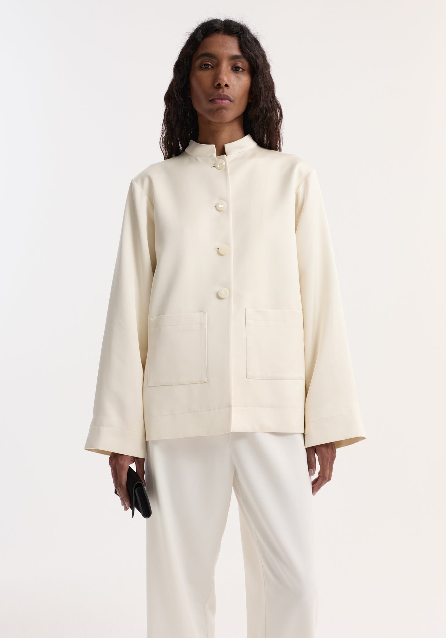 Model in a Pearl button satin jacket | cream with stand-up collar, pearl buttons, and patch pockets, paired with loose white trousers, holding a black clutch.