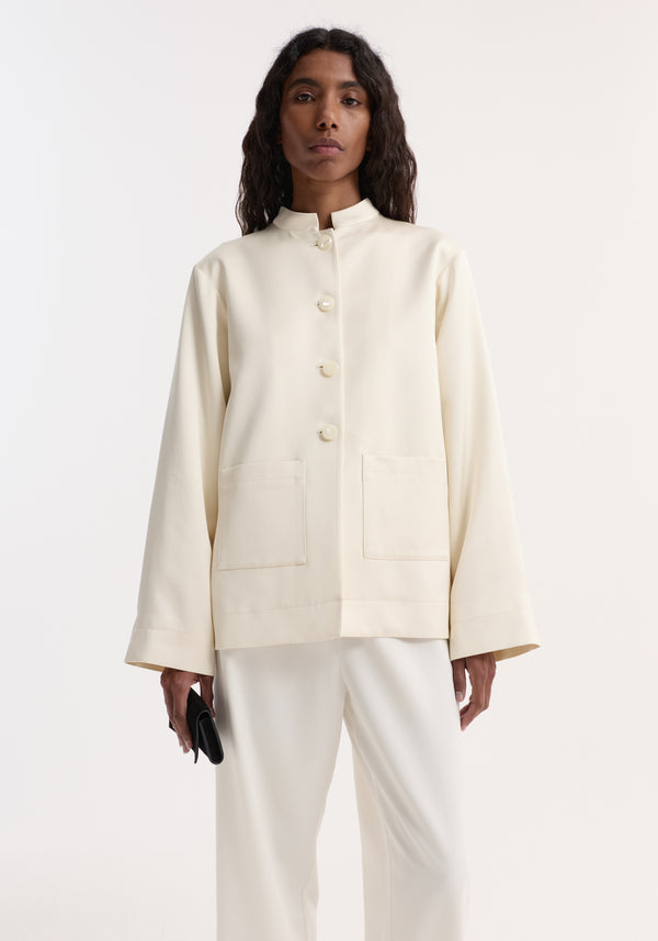 Model in a Pearl button satin jacket | cream with stand-up collar, pearl buttons, and patch pockets, paired with loose white trousers, holding a black clutch.