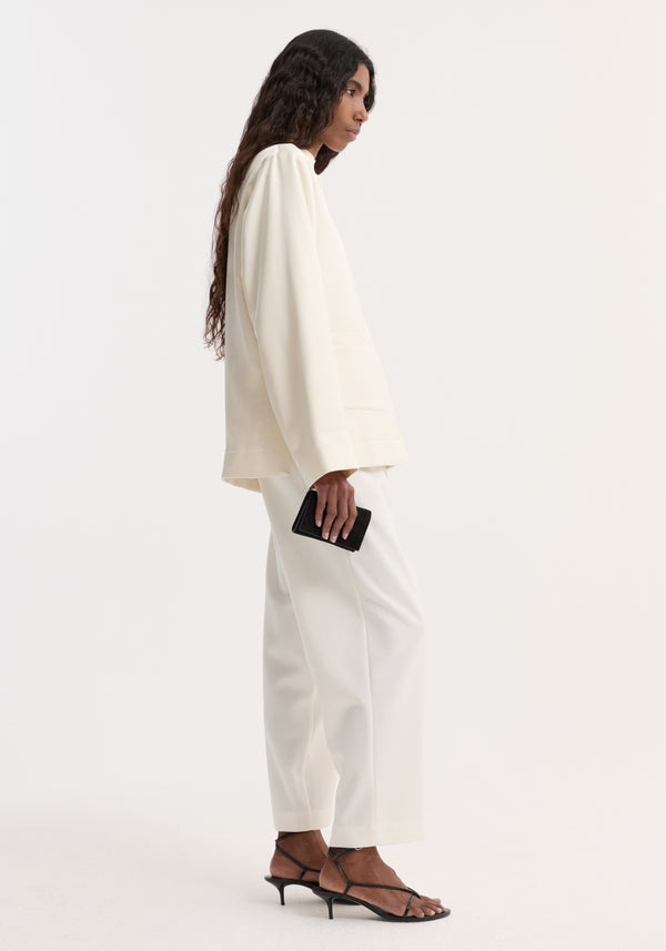 Person in profile wearing Pearl button satin jacket with stand-up collar, pearl buttons, front patch pockets, paired with matching trousers, holding a small black clutch.