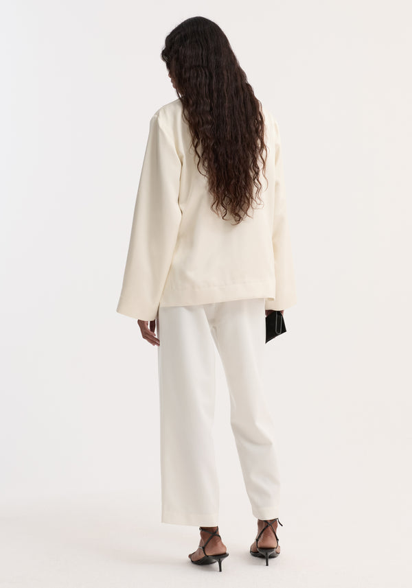 Person wearing Pearl button satin jacket with stand-up collar, pearl buttons, front patch pockets, paired with matching wide-leg trousers, holding a black clutch.