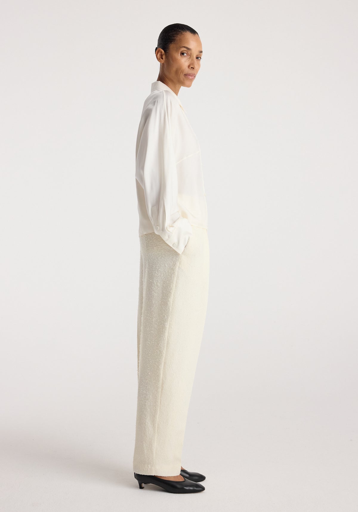 Model wearing Double breasted silk top | cream with long sleeves, paired with cream trousers and black pointed shoes in a minimalist studio setting