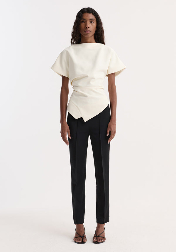 Model in Draped Lapel Top | off white, featuring a slashed neckline and asymmetric peplum, paired with slim black trousers and strappy heels.