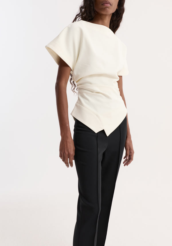 Woman wearing Draped lapel top in off white with bateau neckline, short sleeves, asymmetrical hem, paired with black tailored trousers, highlighting a sculptural silhouette.