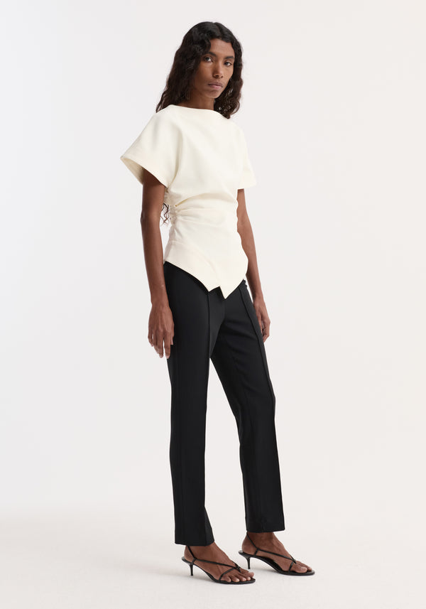 Model wearing the Draped lapel top in off white, showcasing its asymmetrical hem and sculptural design, paired with black trousers and strappy sandals.