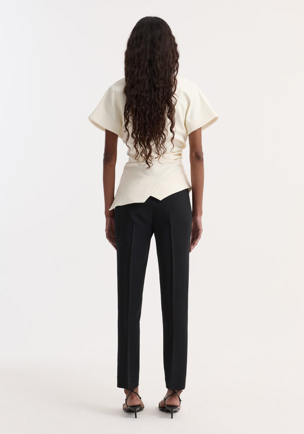 Woman with dark hair wearing Draped lapel top | off white, showcasing an asymmetrical hemline, sculpted shoulder, and reworked lapel, paired with black trousers and sandals.