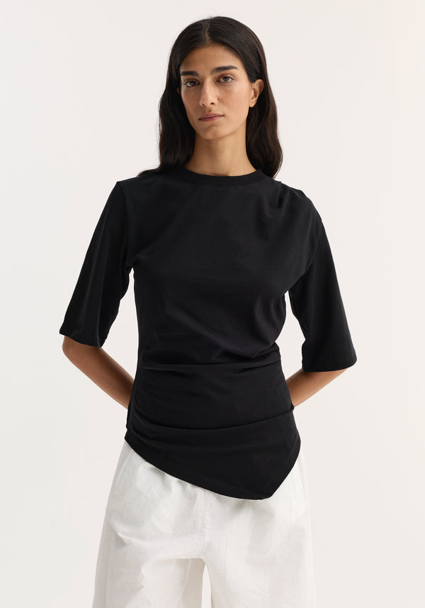 The woman models a Reinvented twisted t-shirt | black, featuring a twisted asymmetrical design and elbow-length sleeves, paired with loose white trousers.