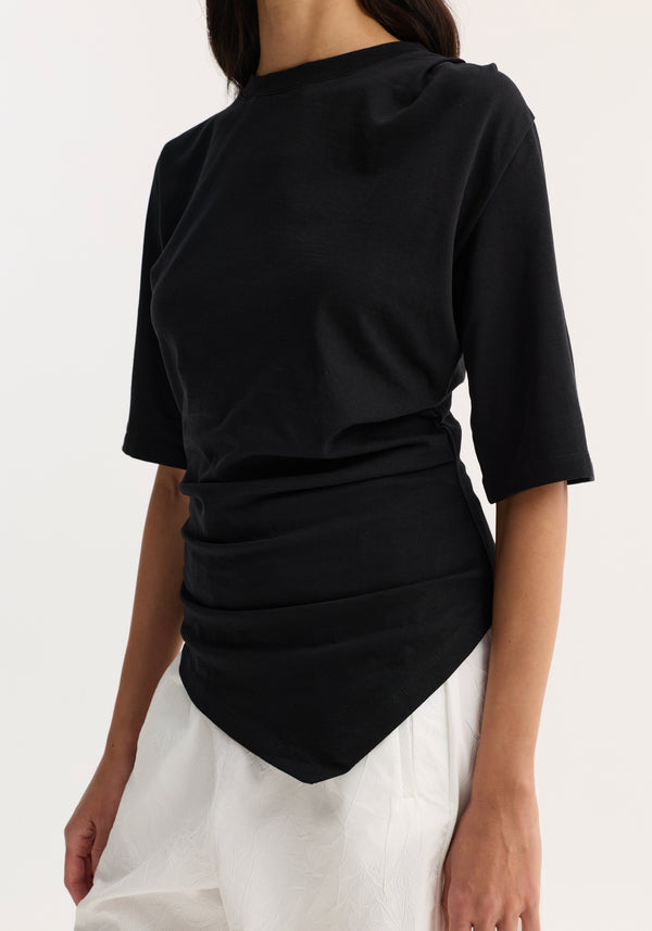 Person wearing a Reinvented twisted t-shirt in black with elbow-length sleeves, ruched waist, and handkerchief hem, paired with loose-fit white trousers.