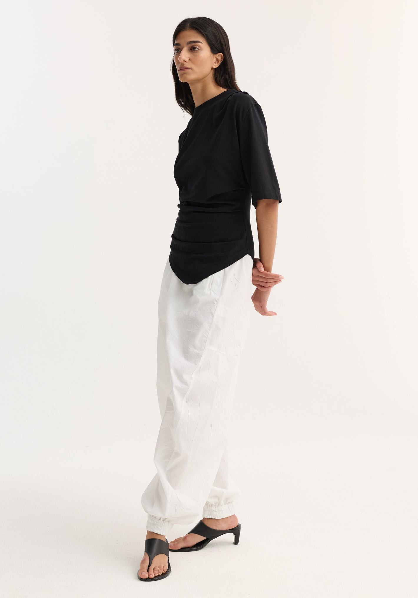 Woman models the Reinvented twisted t-shirt in black, showcasing its asymmetrical draping and fitted waist, paired with white pants and black mules.