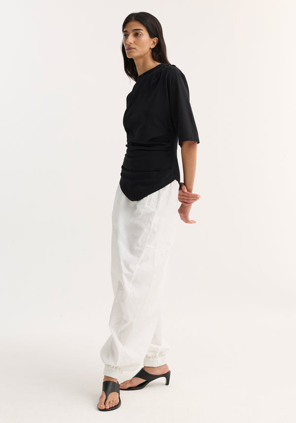 Woman models the Reinvented twisted t-shirt in black, showcasing its asymmetrical draping and fitted waist, paired with white pants and black mules.