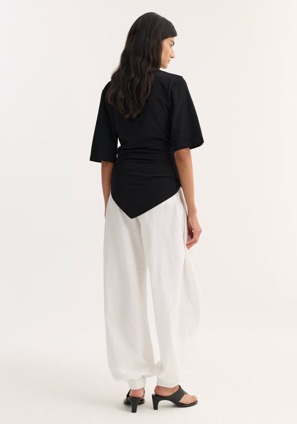 Woman in twisted asymmetrical Reinvented twisted t-shirt, black, showcasing sculptural design with elbow-length sleeves, paired with high-waisted trousers and mid-heel sandals.
