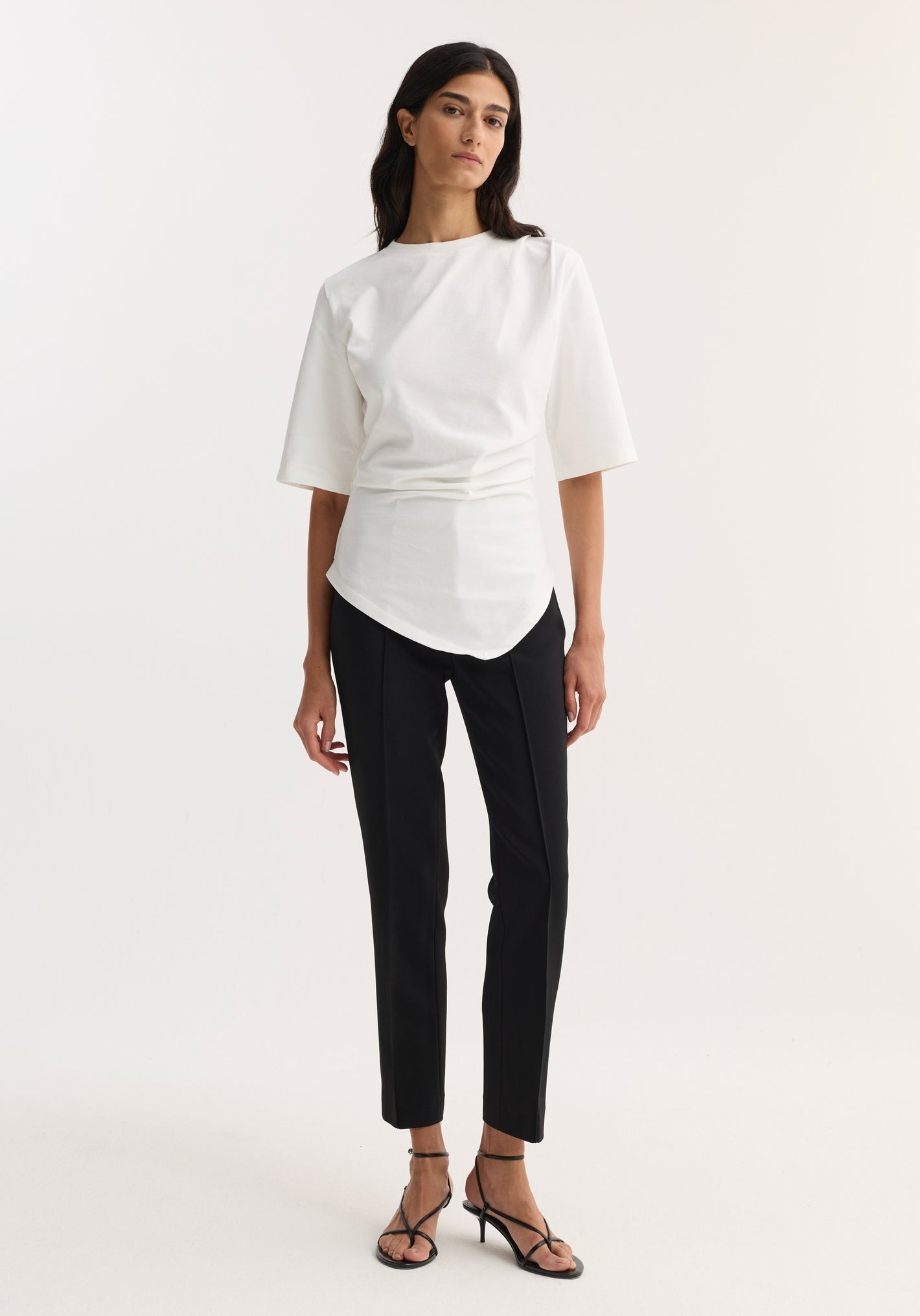 Woman wearing Reinvented twisted t-shirt in optic white, featuring a twisted asymmetrical drape, fitted waist, and elbow-length sleeves, paired with slim-fit black trousers and strappy sandals.