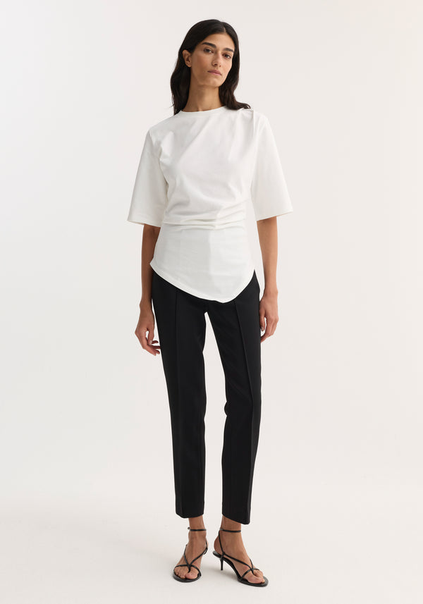 Woman wearing Reinvented twisted t-shirt in optic white, featuring a twisted asymmetrical drape, fitted waist, and elbow-length sleeves, paired with slim-fit black trousers and strappy sandals.