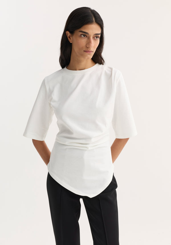 Woman wearing Reinvented twisted t-shirt in optic white with elbow-length sleeves and twisted asymmetrical design, paired with black tailored trousers, standing against a light backdrop.