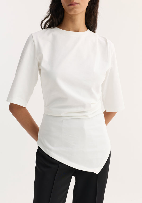 Woman wearing the Reinvented twisted t-shirt in optic white, featuring an asymmetrical hem and elbow-length sleeves, paired with straight-leg black trousers.