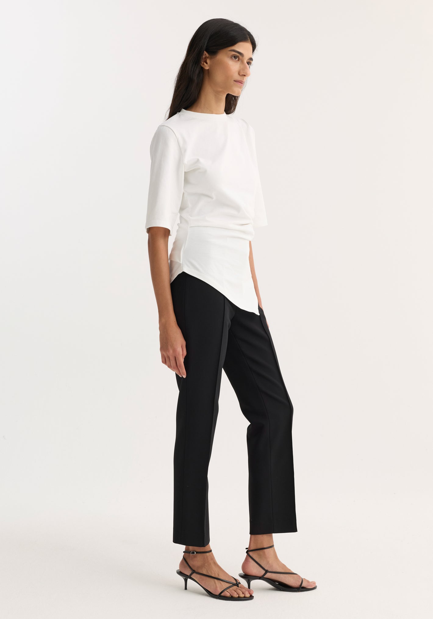 Woman modeling a Reinvented twisted t-shirt in optic white with elbow-length sleeves, featuring a twisted asymmetrical design, paired with black trousers and strappy sandals.