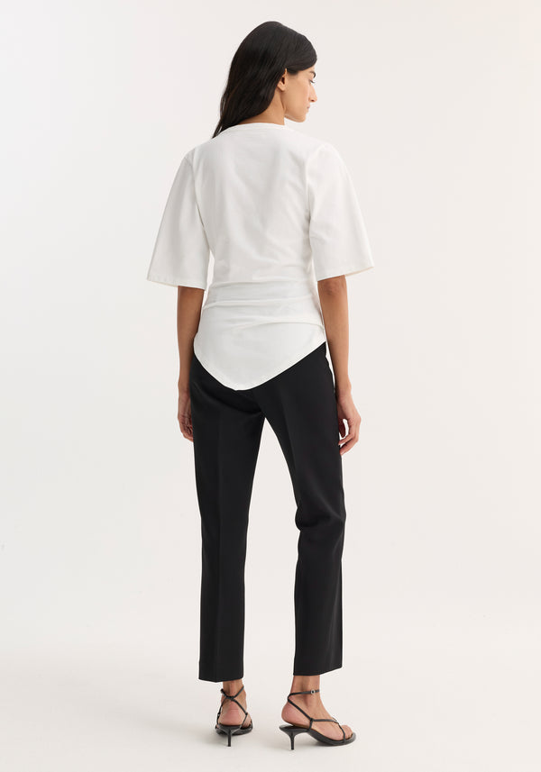 Woman models Reinvented twisted t-shirt in optic white, showcasing its asymmetrical drape and fitted waist, paired with slim black trousers and strappy heels.